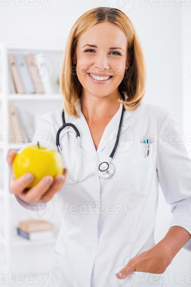 Eat healthy Cheerful female doctor in white uniform stretching out hand with green apple and smiling photo