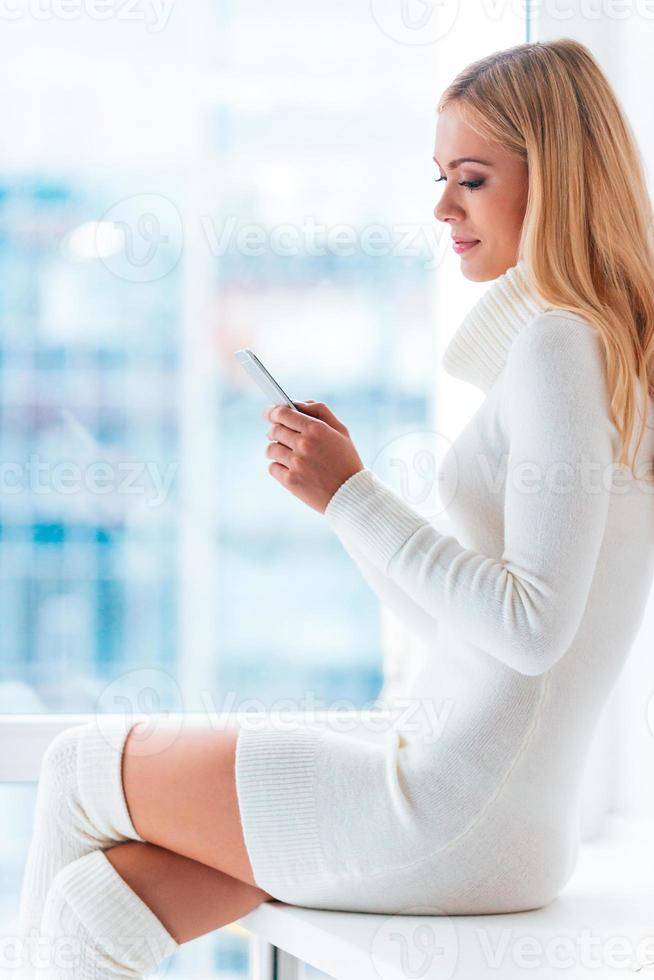 Reading message from him. Side view of beautiful young woman in warm sweater and socks holding smart phone and looking at it while sitting in front of the window at home photo