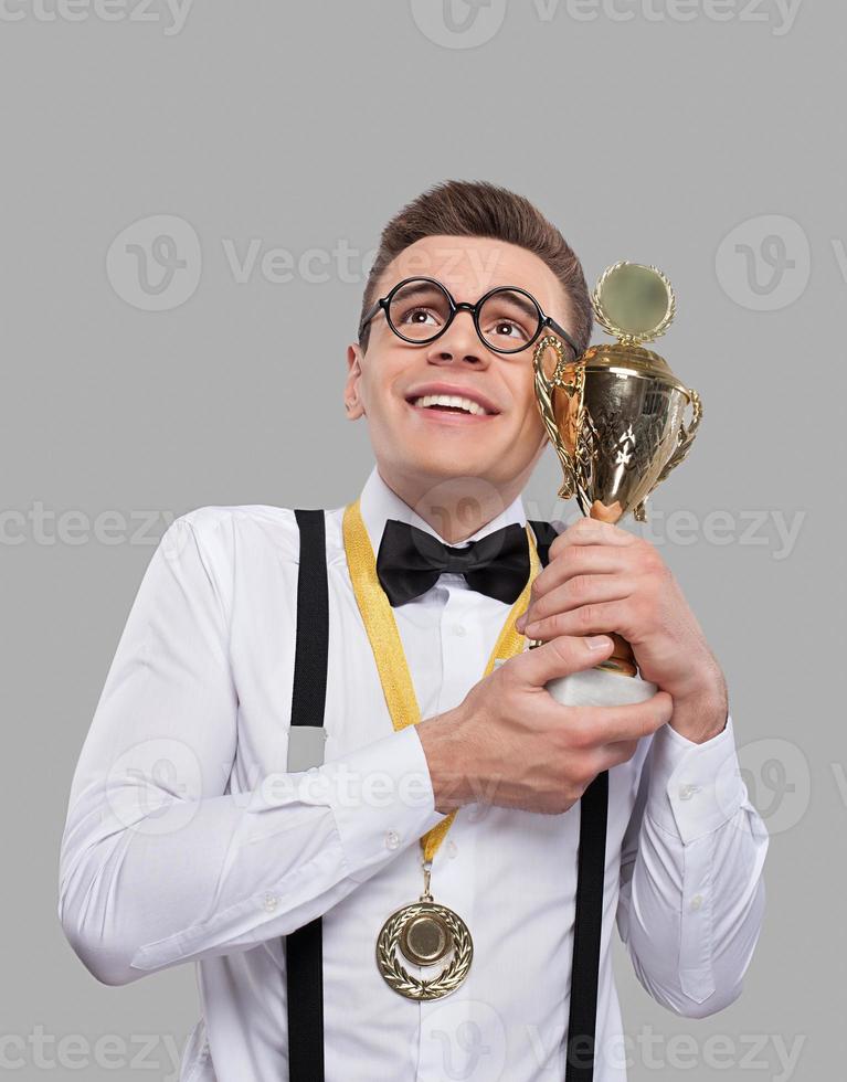 soy el mejor joven alegre con corbata de moño sosteniendo un trofeo y mirando hacia arriba mientras estoy de pie contra un fondo gris foto