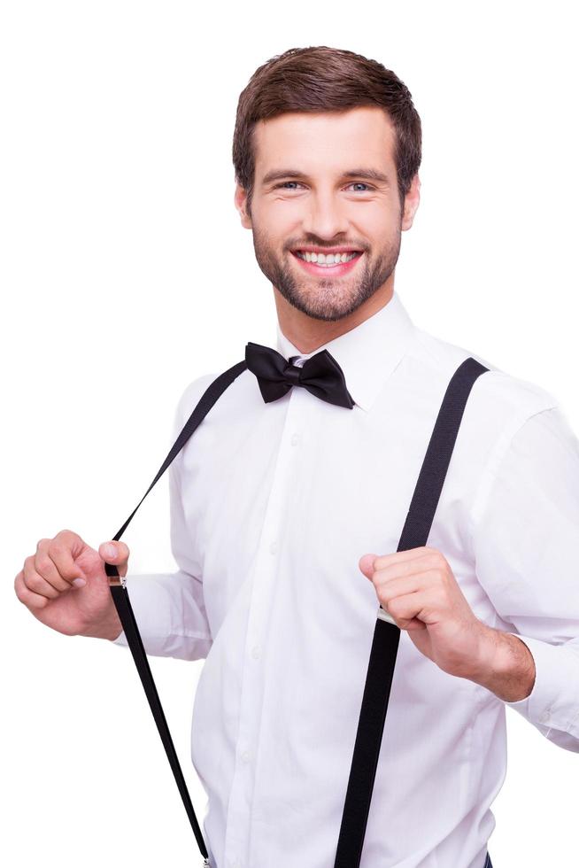 novio feliz retrato de un joven apuesto con camisa blanca y corbata de moño ajustando sus tirantes y sonriendo a la cámara mientras está de pie aislado en blanco foto