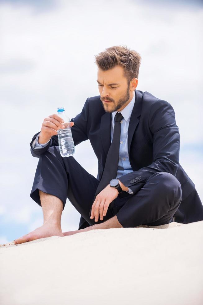 Water is life. Depressed young businessman holding bottle with water and looking at it while sitting on sand photo