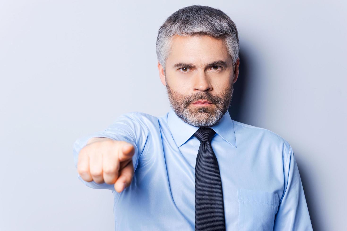 I made my choice. Confident mature man in shirt and tie looking at camera and pointing you while standing against grey background photo