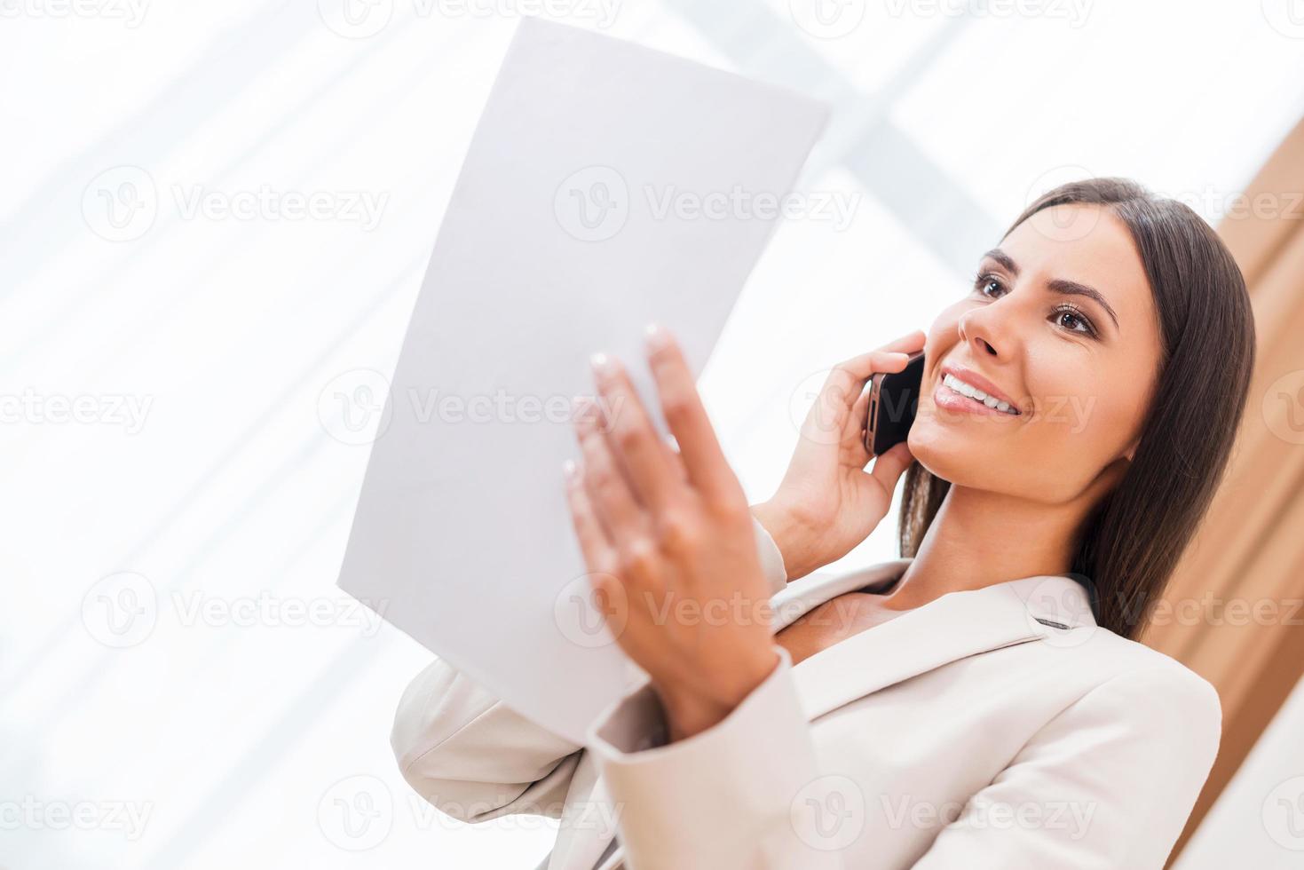 Happy with a contract signed. Low angle view of happy young businesswoman in suit talking on the mobile phone and holding document photo