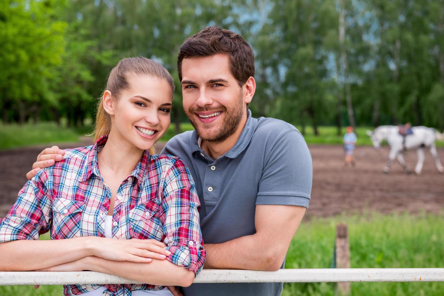 Couple on ranch. Happy young loving couple standing close to each other and smiling while standing on ranch with horse walking in the background photo