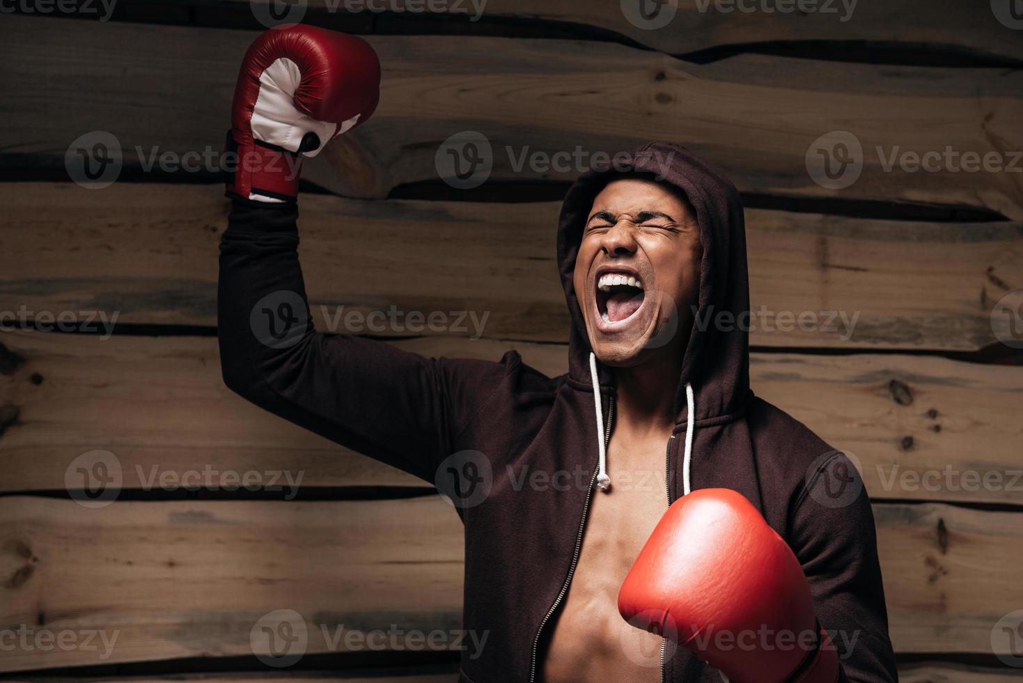 solía ganar. feliz joven africano con camisa encapuchada y guantes de boxeo levantando los brazos y manteniendo los ojos cerrados mientras está de pie contra un fondo de madera foto