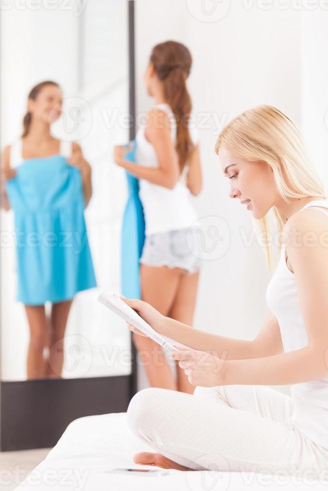 How this dress fits me Beautiful young woman holding dress and looking at the mirror while another women sitting on the foreground and reading magazine photo