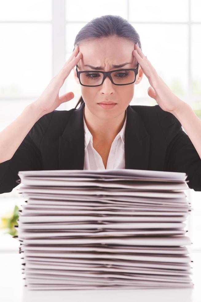 That is too much. Depressed young woman in suit tie looking at the stack of paperwork and holding head in hands while sitting at her working place photo