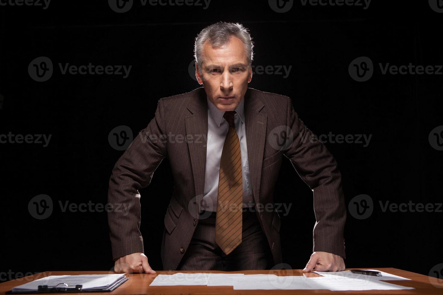 soy un jefe aquí un hombre mayor serio en ropa formal mirando a la cámara mientras se apoya en la mesa y aislado en negro foto