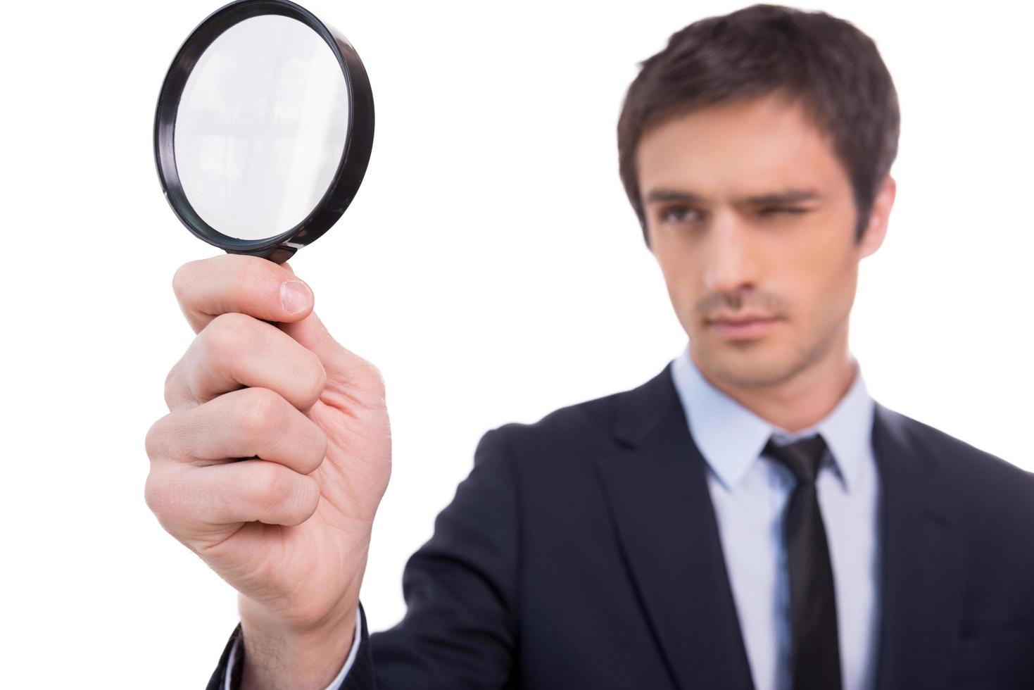 Looking through magnifying glass. Concentrated young man in formalwear looking through a magnifying glass while standing isolated on white background photo