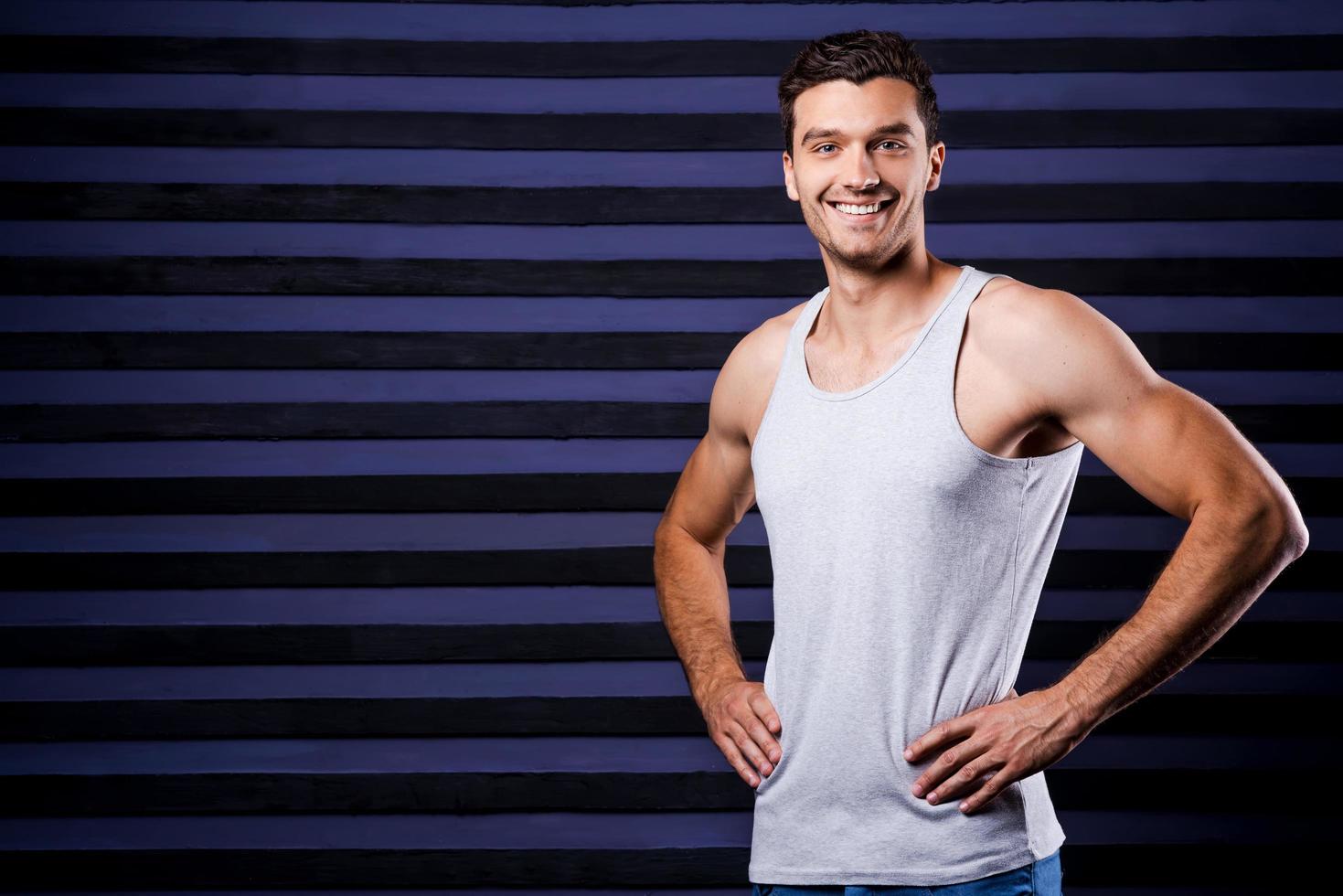 deportivo y guapo. joven deportivo en la parte superior del tanque cogido de la mano en la cadera y sonriendo mientras está de pie contra el fondo rayado foto