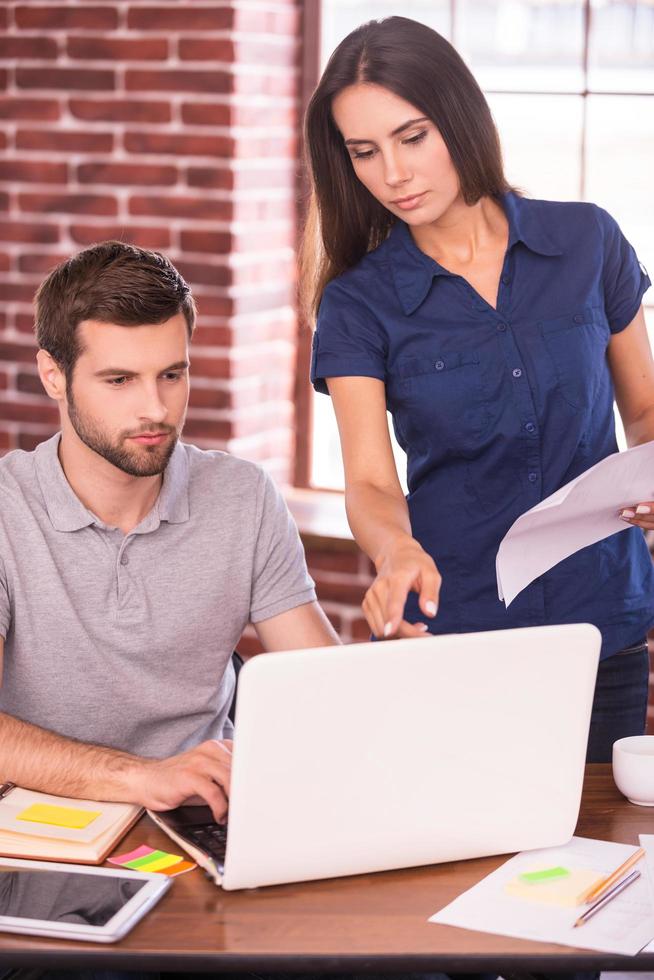 consultando con un colega. joven seguro de sí mismo sentado en su lugar de trabajo y mirando la computadora portátil mientras una mujer hermosa se para cerca de él y señala el monitor foto