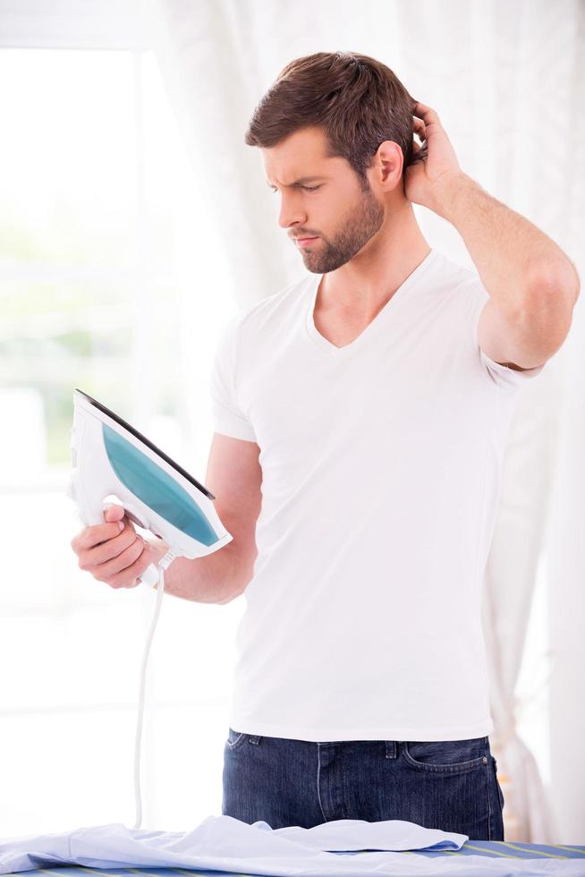 Something wrong with this iron. Frustrated young man holding iron and looking at it while standing near the ironing board at home photo