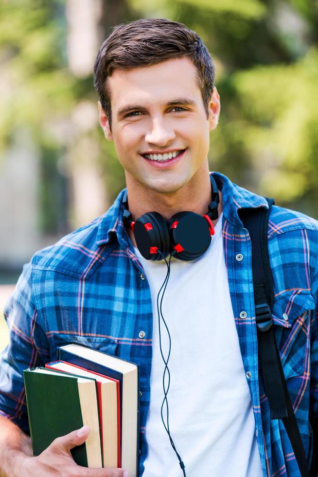 Confident and successful student. Handsome young man holding books and smiling while standing outdoors photo
