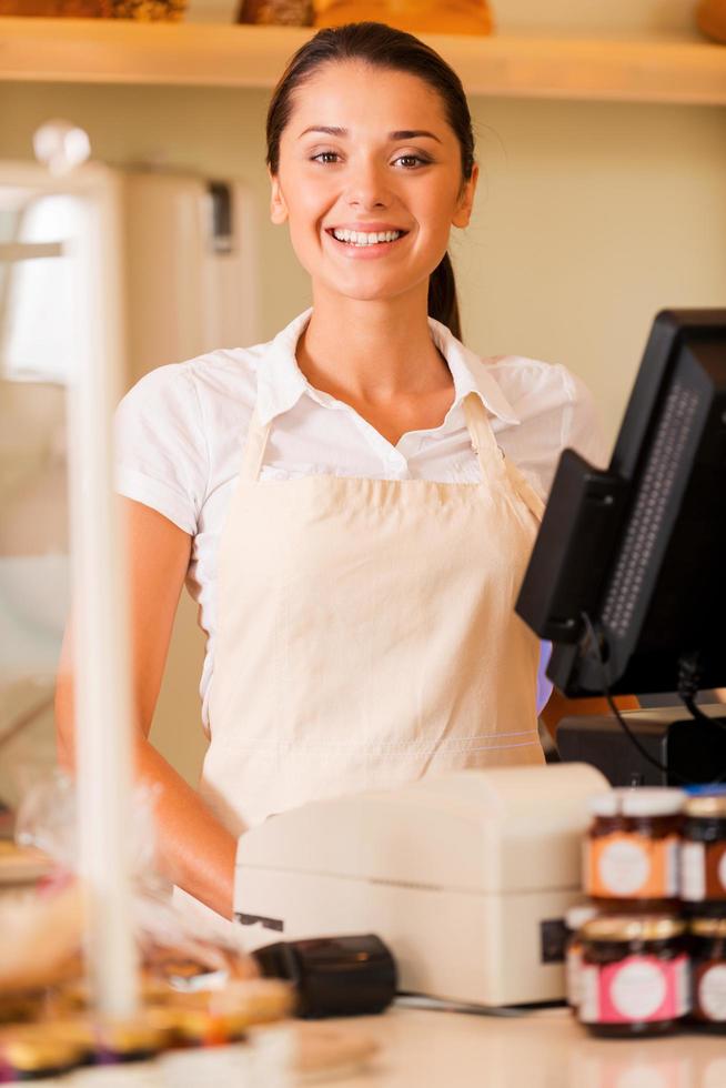 Cheerful cashier. Beautiful young female cashier in apron standing near cash register and smiling photo