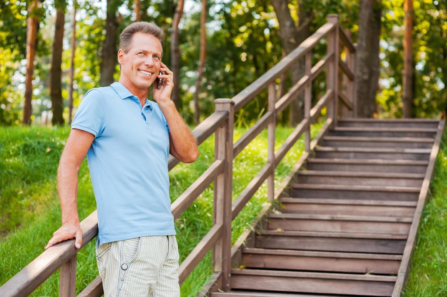 Good talk with friend. Cheerful mature man talking on the mobile phone and smiling while standing at wooden staircase photo