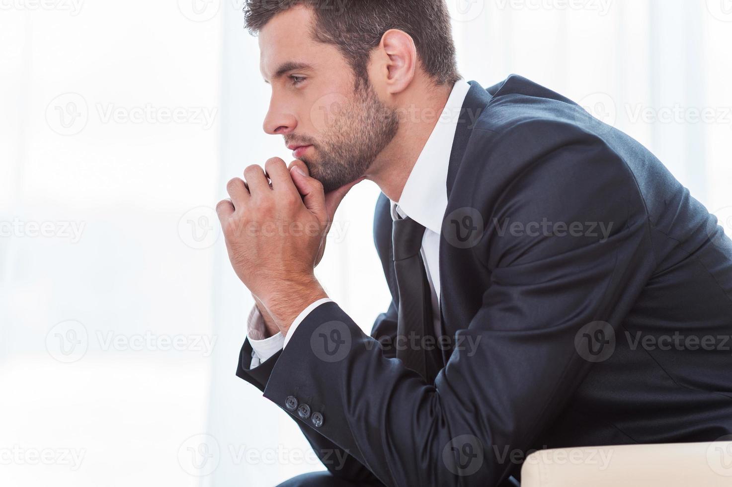 Thoughtful businessman. Side view of thoughtful young businessman in shirt and tie holding hands on chin and looking away while sitting at the chair photo