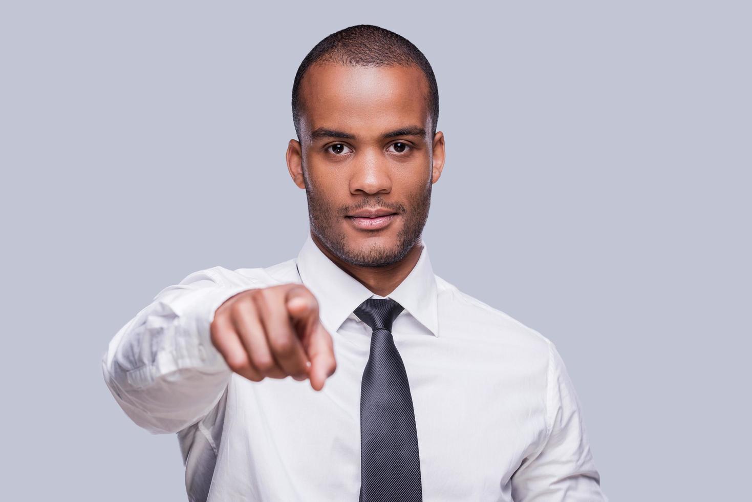 I choose you. Confident young African man in shirt and tie pointing you while standing against grey background photo