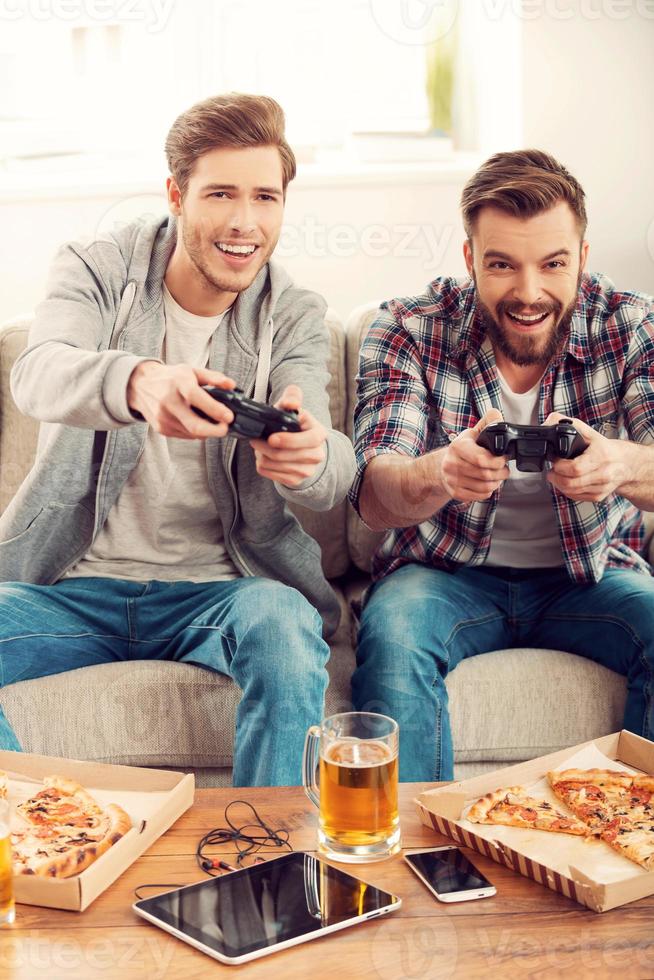 We love this game Two cheerful young men playing video games while sitting on sofa photo