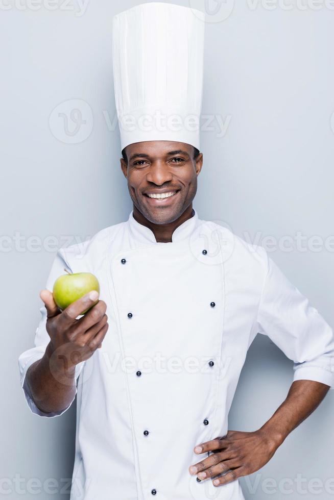 un ingrediente importante para la ensalada de frutas. confiado joven chef africano con uniforme blanco estirando manzana verde y mirando la cámara con una sonrisa mientras se enfrenta a un fondo gris foto