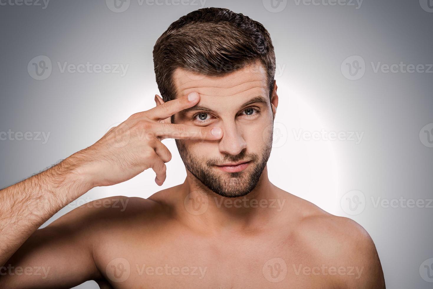 My eyes on you Portrait of handsome young shirtless man looking at camera and holding fingers near his eye while standing against grey background photo