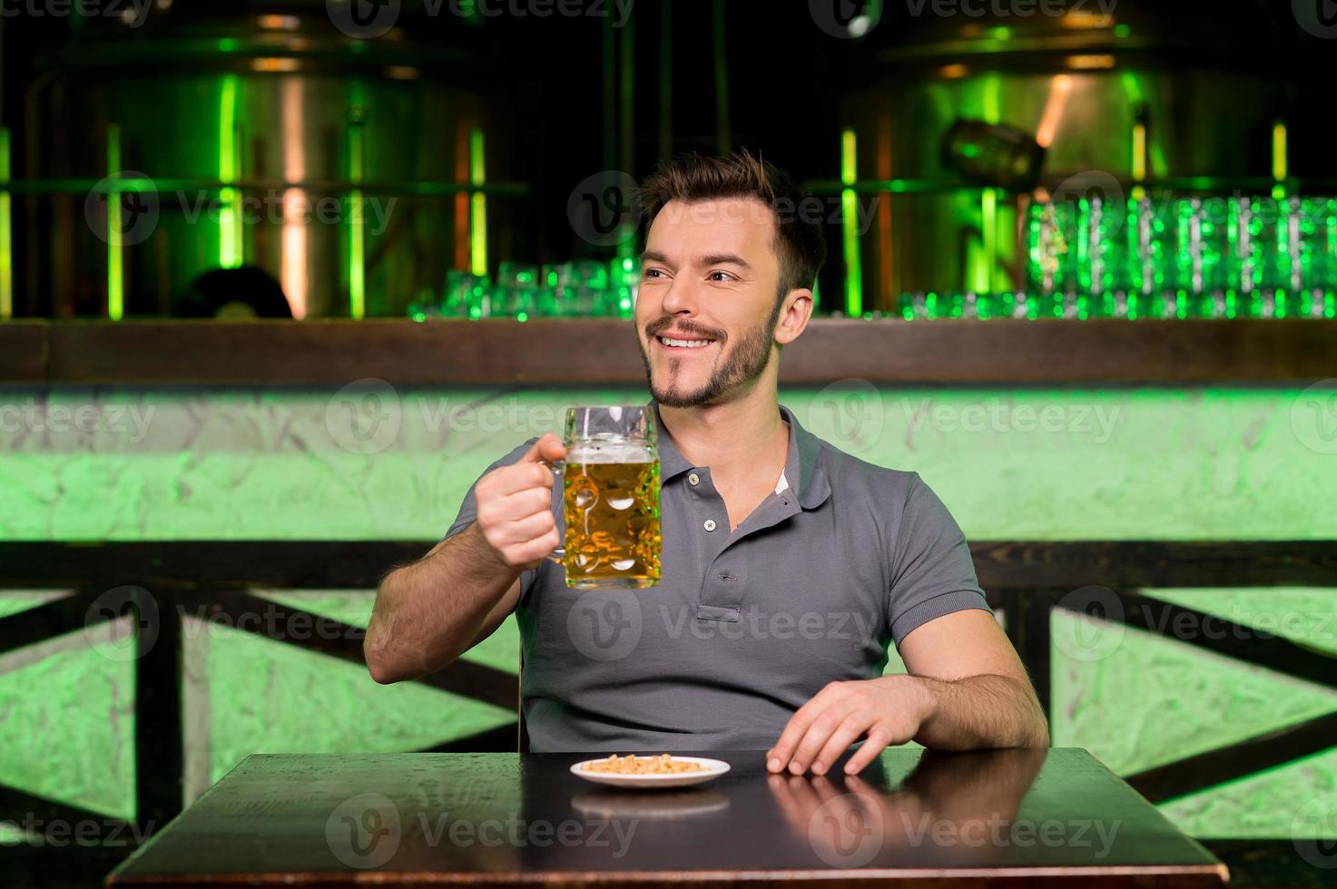 What a great day Cheerful young man holding a mug with beer and smiling while sitting in bar photo