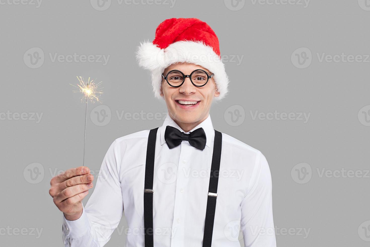 feliz año nuevo alegre joven nerd con corbata de moño y tirantes sosteniendo una bengala y sonriendo mientras se enfrenta a un fondo gris foto