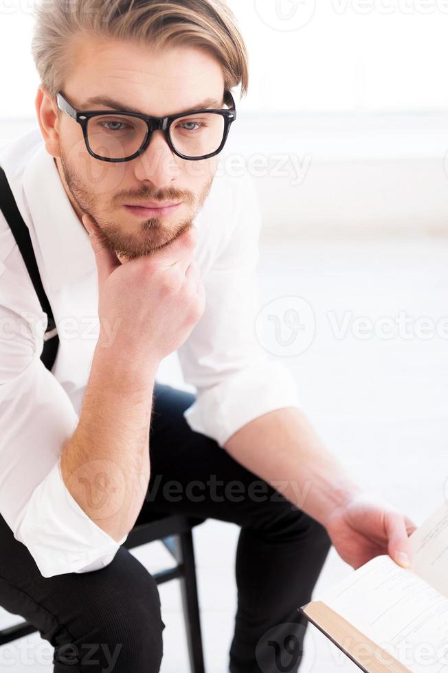 What an interesting fact Top view of thoughtful young man in shirt and suspenders holding a book and looking away while sitting on the chair photo