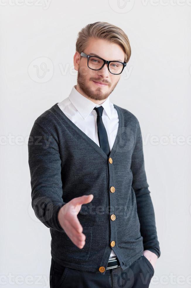 Welcome on board Handsome young man in glasses stretching out hand for shaking while standing against grey background photo