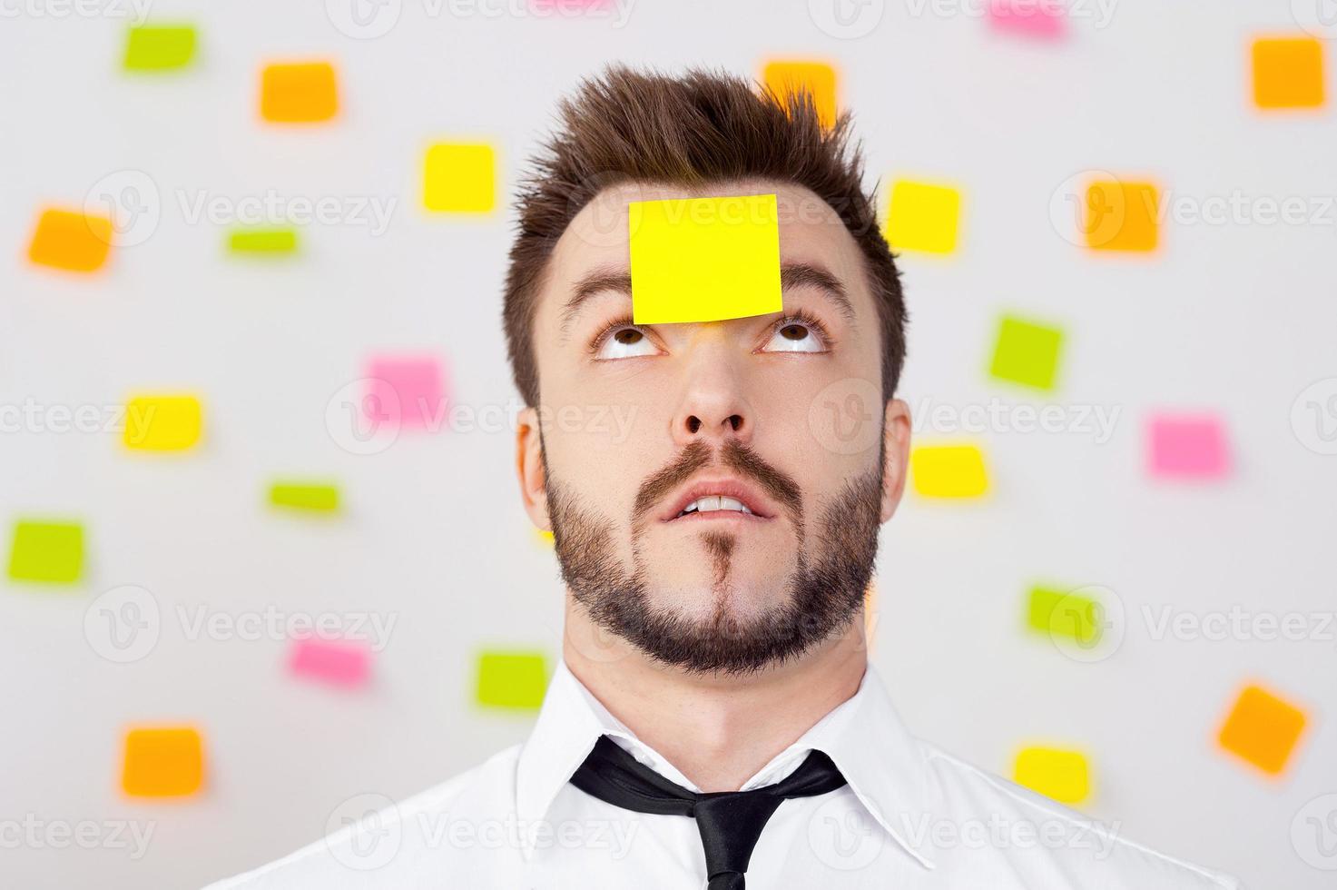 So much work to do Portrait of frustrated young man in formalwear and adhesive note on his forehead standing against the wall with many adhesive notes on it photo