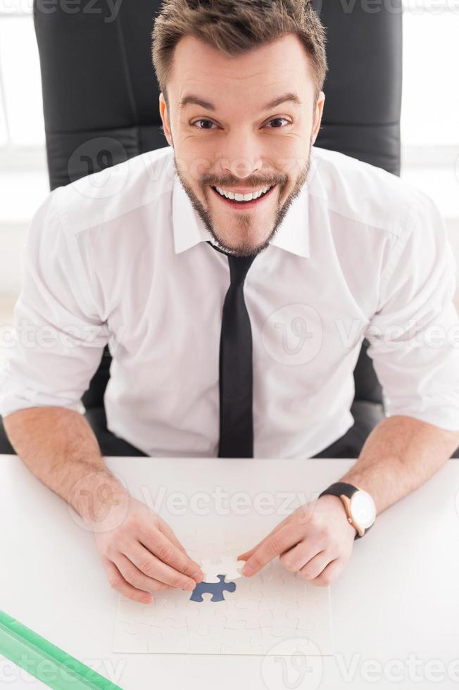 I did it Top view of happy young man in shirt and tie collecting puzzle while sitting at his working place photo