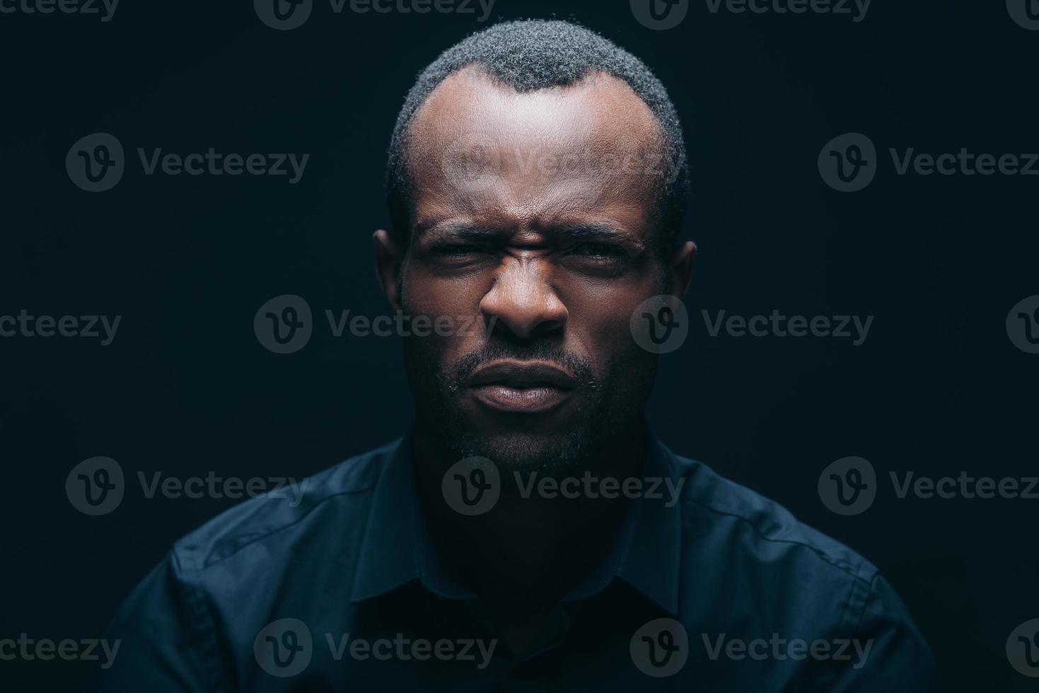 este es un retrato repugnante de un joven africano mirando a la cámara y expresando disgusto mientras está frente a un fondo negro foto