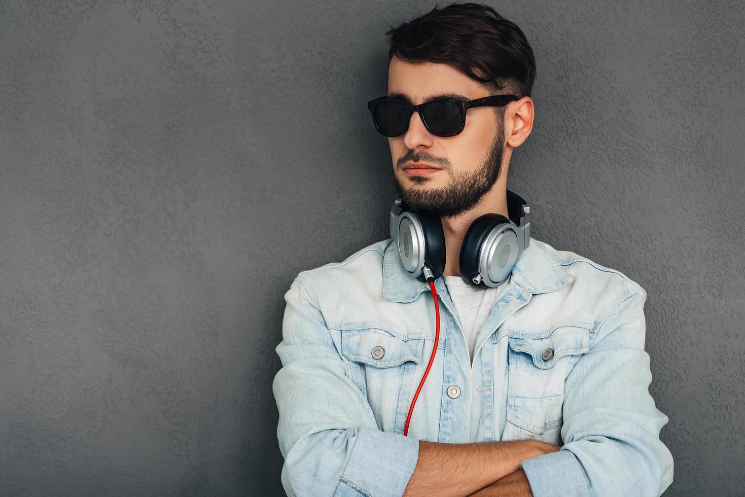 seguro y elegante. joven en auriculares con los brazos cruzados y mirando hacia otro lado mientras está de pie contra el fondo gris foto