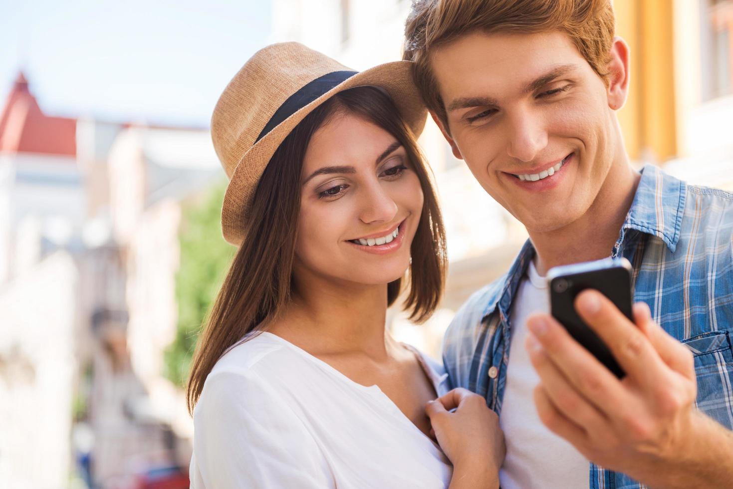 Sharing their pictures with friends. Beautiful young loving couple standing close to each other while man holding mobile phone and smiling photo
