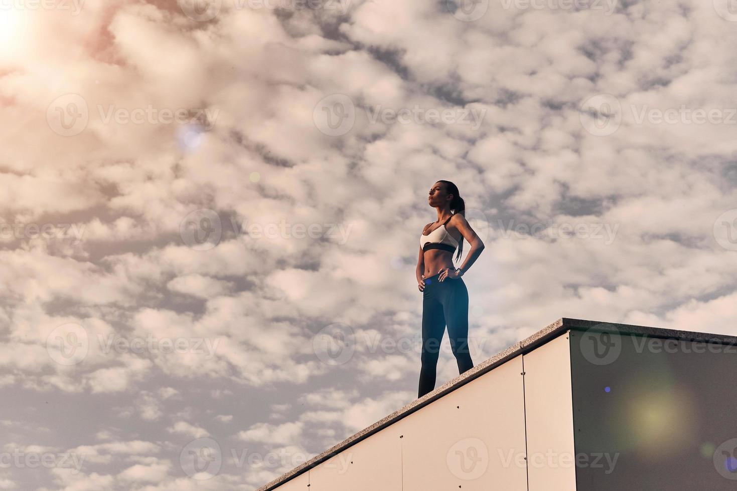 su cuerpo se ganó un descanso. mujer joven moderna en ropa deportiva relajándose mientras está de pie en el techo al aire libre foto