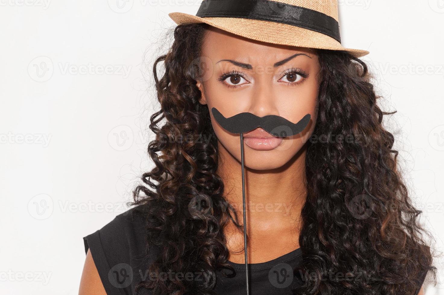 What do you think about my mustache Portrait of playful young African woman in funky hat holding fake mustache on her face and looking at camera while standing against white background photo