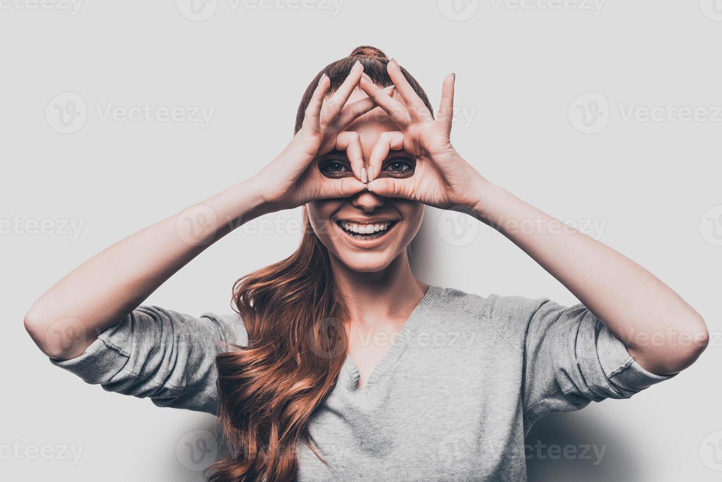 Just for fun. Joyful young woman gesturing eyeglasses near face and smiling while standing against grey background photo