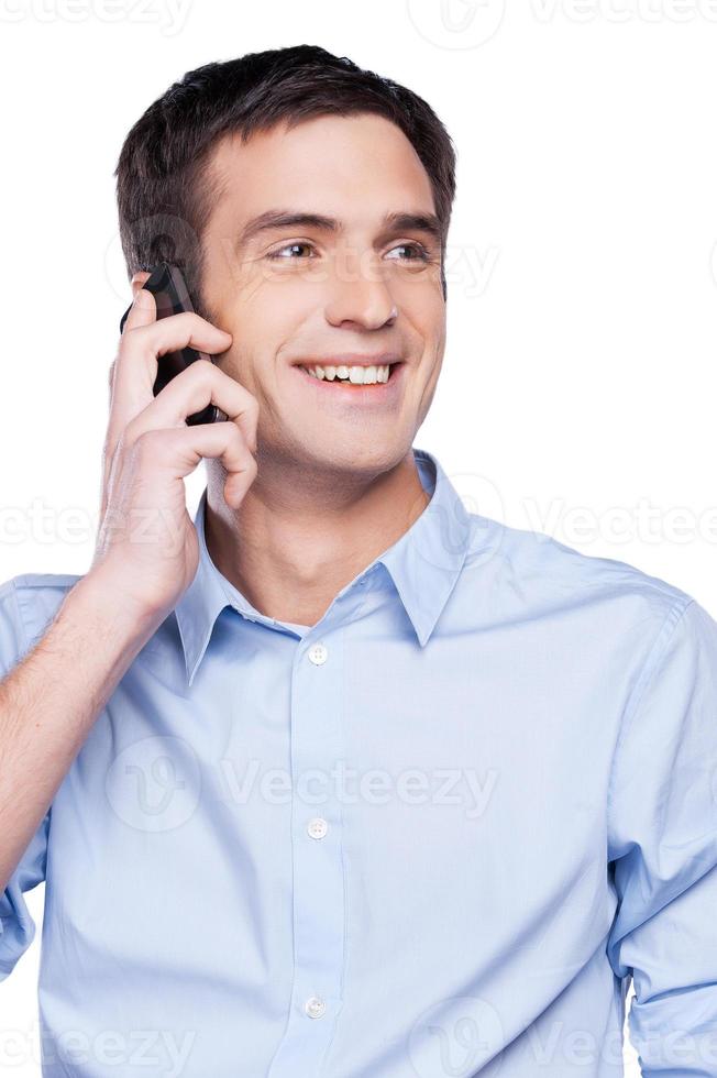 buenas noticias un joven apuesto con camisa azul hablando por teléfono móvil y sonriendo mientras está aislado en blanco foto