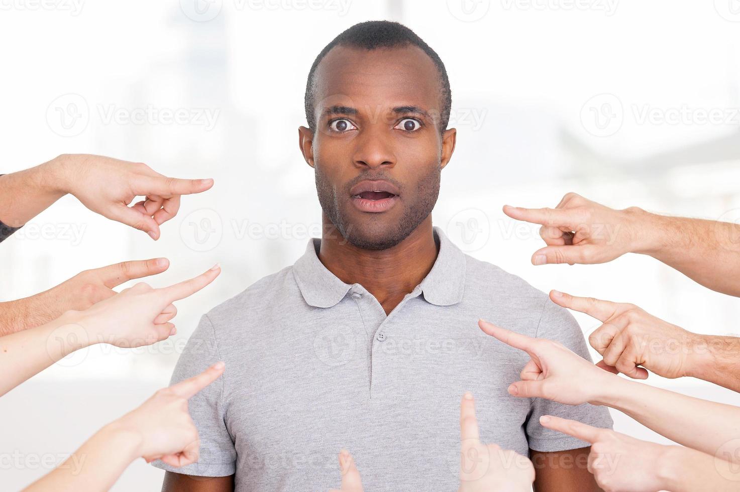 Why me Shocked young African man looking at camera while many hands pointing him photo