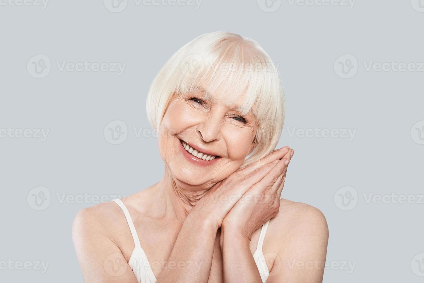 Aging process. Beautiful senior woman looking at camera and smiling while standing against grey background photo