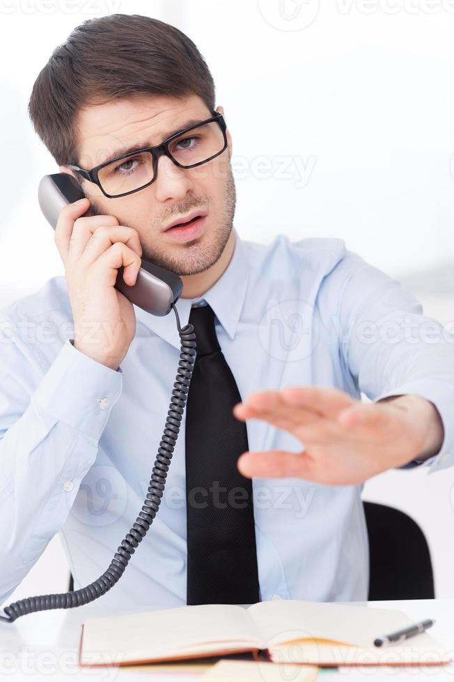 Not now Angry young man in shirt and tie talking on the mobile phone and gesturing while sitting at his working place photo