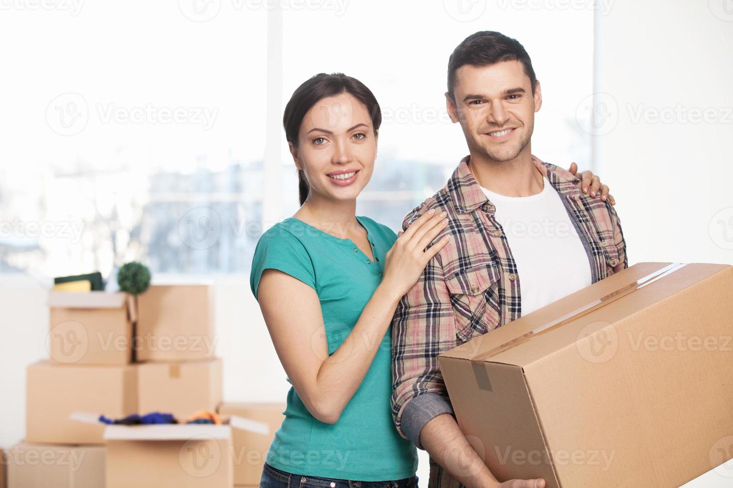 Moving to a new house. Cheerful young couple standing close to each other and smiling at camera while man holding a cardboard box photo