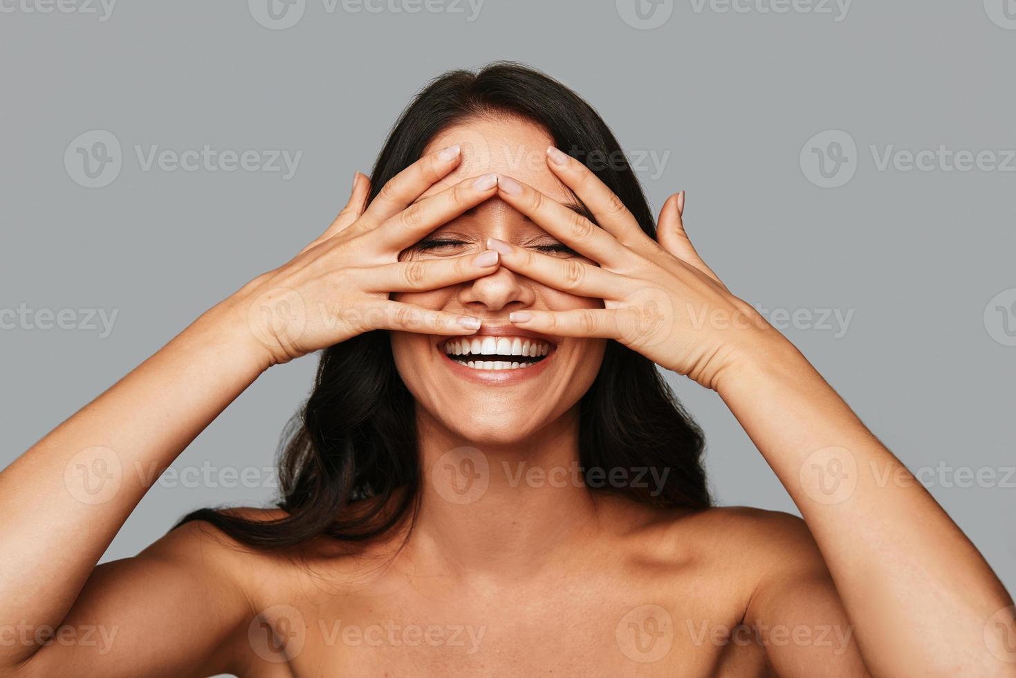 Feeling playful. Beautiful young woman keeping eyes closed and covering face with hands while standing against grey background photo