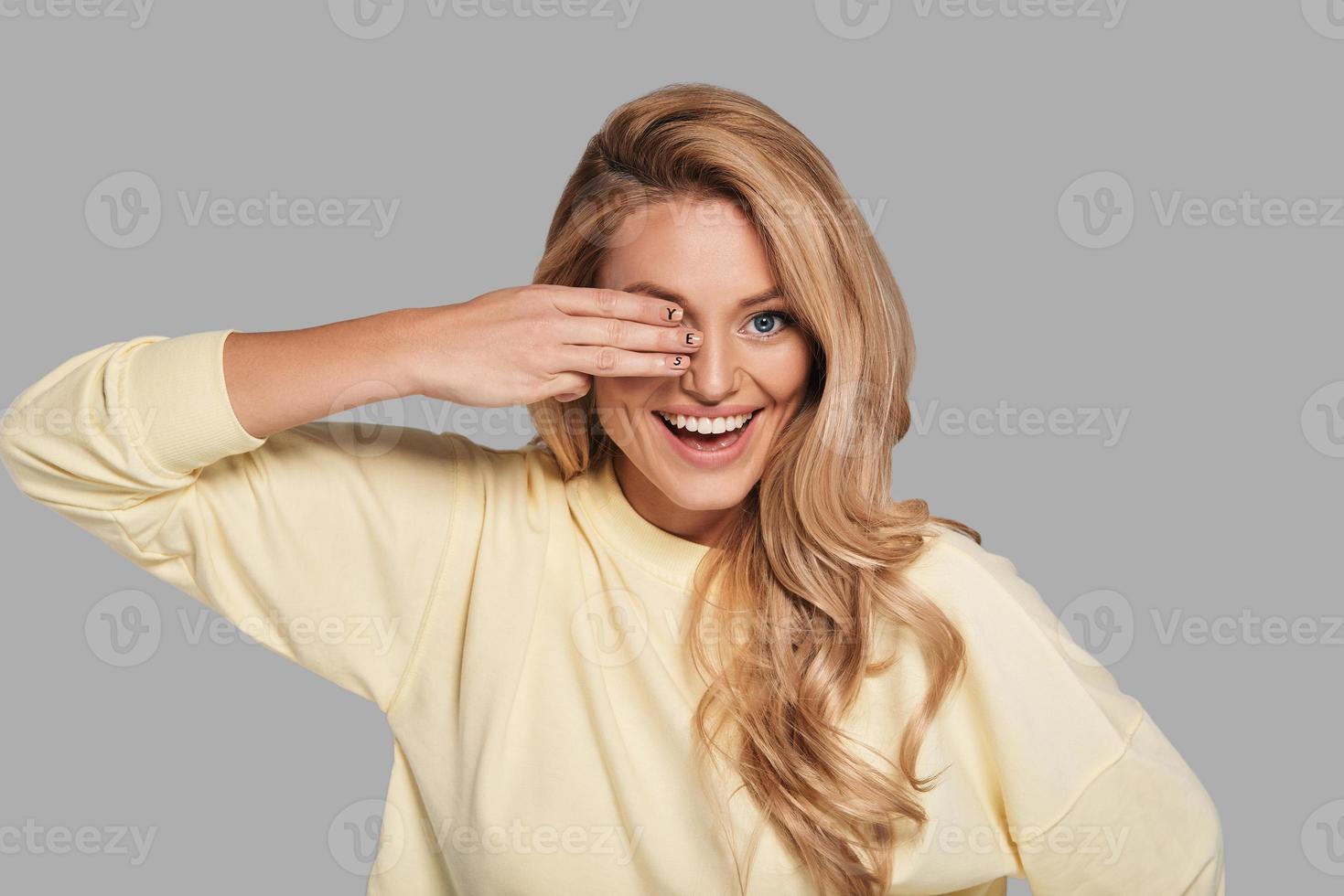Never give up  Attractive young smiling woman covering eye with hand and looking at camera while standing against grey background photo