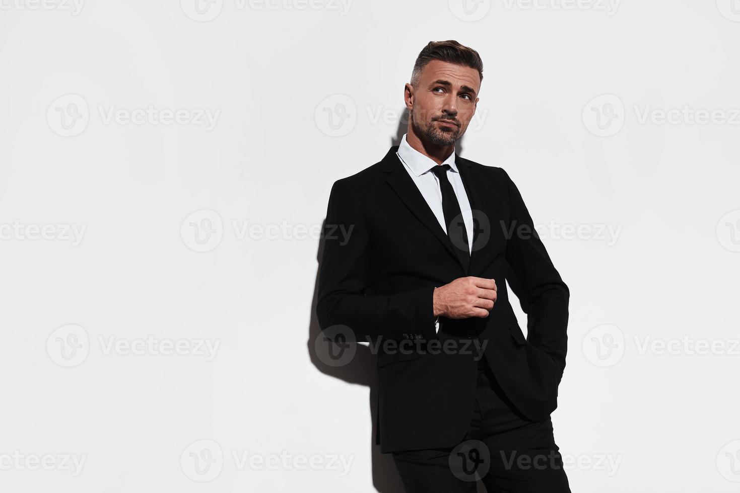 Thinking about... Handsome young man in full suit keeping hand in pocket and looking away while standing against white background photo