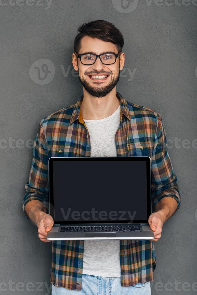 Pay attention to this Confident young man showing his laptop and looking at camera with smile while standing against grey background photo