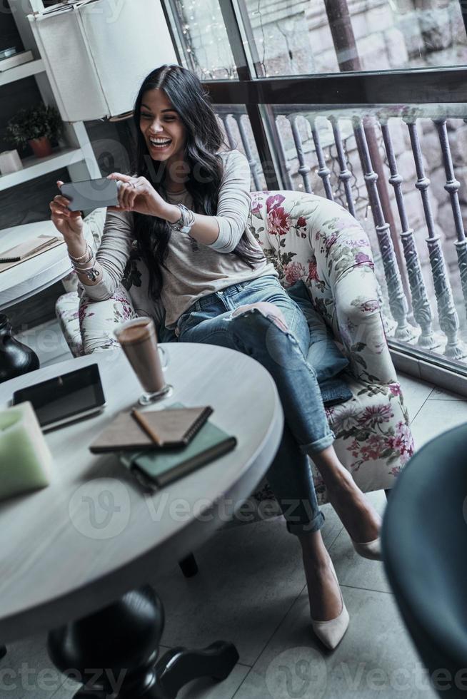 Sharing self portraits with her boyfriend. Attractive young smiling woman taking selfie using her smart phone while sitting in restaurant photo