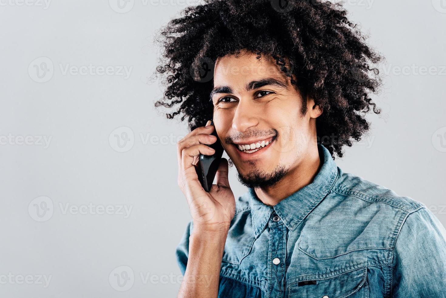 Great phone call. Portrait of handsome young African man talking on mobile phone and looking away with smile while standing against grey background photo