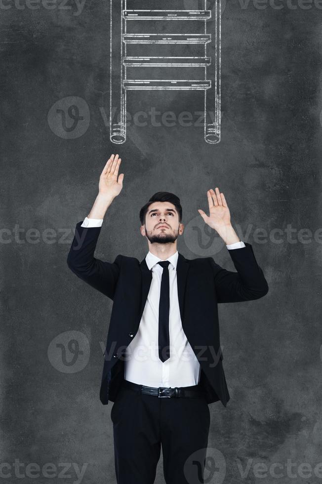 Helping ledder. Handsome young man in formalwear stretching out his hands and looking up while standing against chalkboard with drawing ledder on it photo
