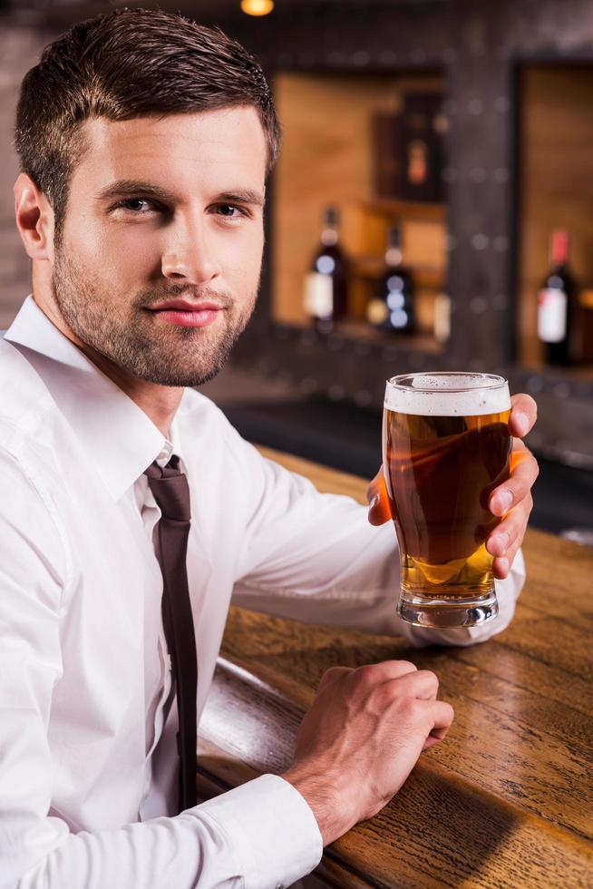 Refreshing with glass of cold beer. Side view of handsome young man in shirt and tie holding glass with beer and looking at camera while sitting at the bar counter photo