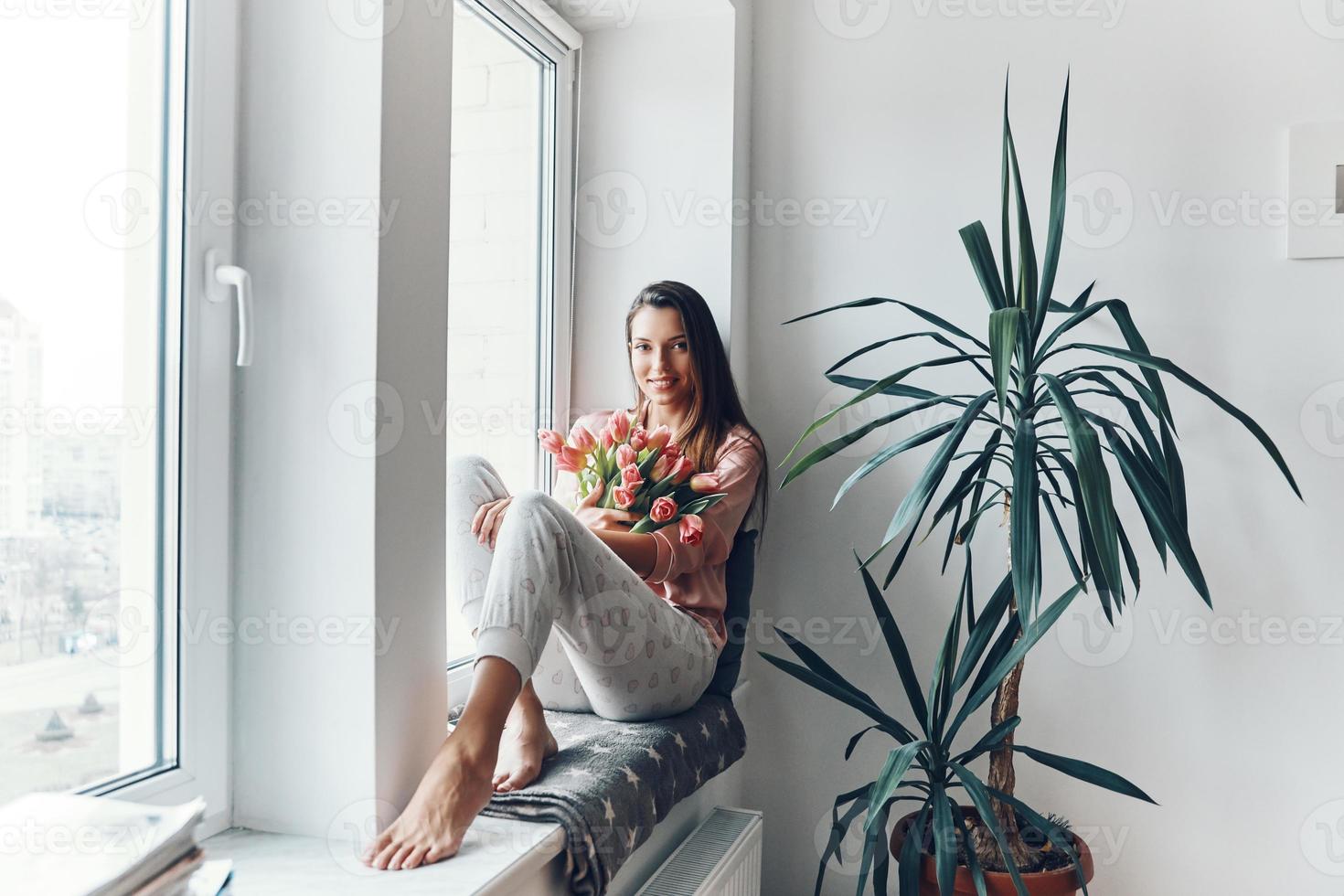 mujer joven atractiva en pijamas acogedores disfrutando de su ramo de tulipanes y mirando a la cámara mientras descansa en el alféizar de la ventana en casa foto